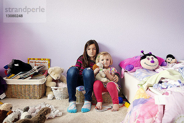 Porträt von Schwestern  die im Schlafzimmer inmitten von Spielzeug an der Wand sitzen