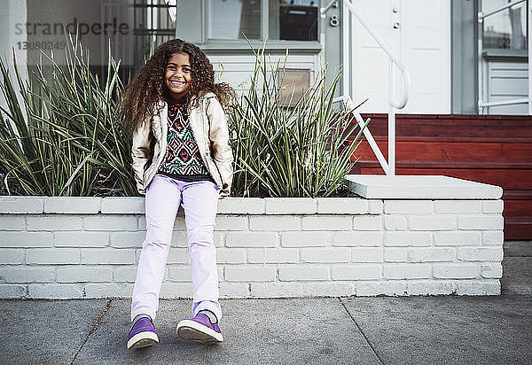 Porträt eines glücklichen Mädchens  das auf einer Stützmauer am Bürgersteig sitzt