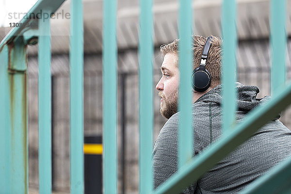 Seitenansicht eines nachdenklichen Mannes  der über Kopfhörer Musik hört  durch ein Geländer in der Stadt gesehen