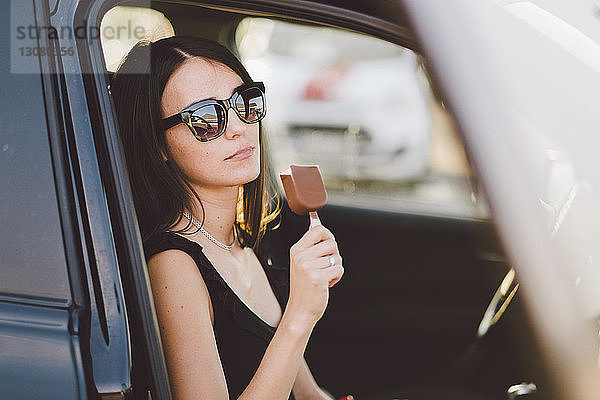 Junge Frau mit Sonnenbrille hält gefrorene Süßspeisen in der Hand  während sie im Auto sitzt und durch das Fenster gesehen wird