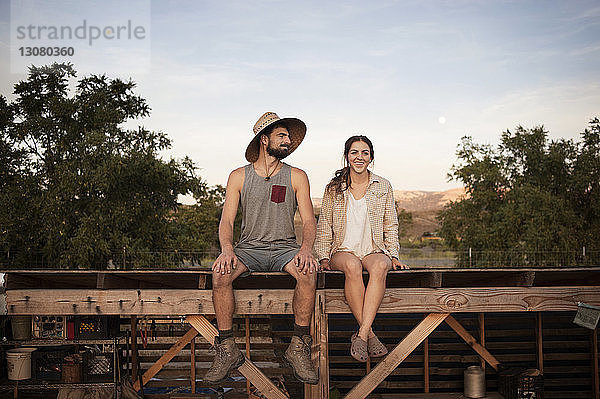 Glückliche Bäuerin sitzt mit Mann auf Holzkonstruktion auf dem Bauernhof