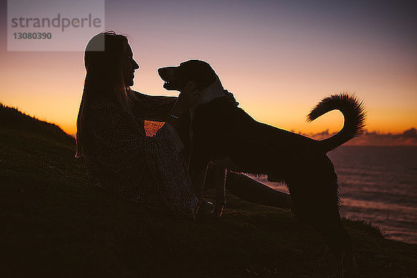 Seitenansicht einer Frau  die bei Sonnenuntergang mit Hund auf einem Hügel gegen den Himmel sitzt