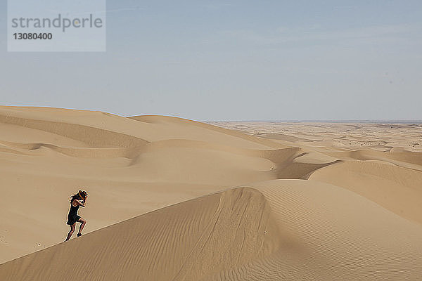 Seitenansicht einer jungen Frau mit Hut beim Klettern auf einer Sanddüne in der Wüste gegen den Himmel