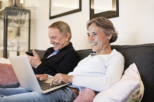 Fröhliches älteres Ehepaar mit Laptop und Smartphone auf dem Sofa zu Hause
