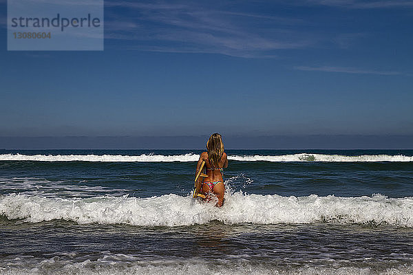 Rückansicht einer Frau  die ein Surfbrett trägt  während sie am sonnigen Tag im Meer vor blauem Himmel steht