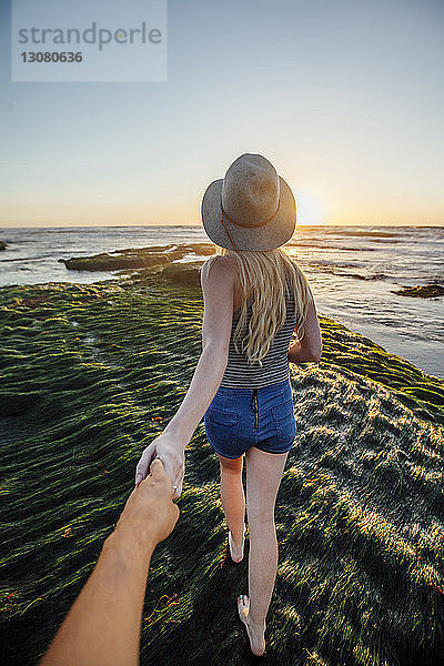 Abgehackte Hand eines Mannes  der die Hand seiner Freundin hält  während er am Strand bei Sonnenuntergang über Gras läuft