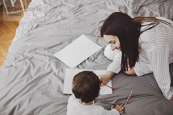 Hochwinkelaufnahme einer glücklichen Mutter und eines kleinen Jungen mit Schulsachen auf dem Bett zu Hause