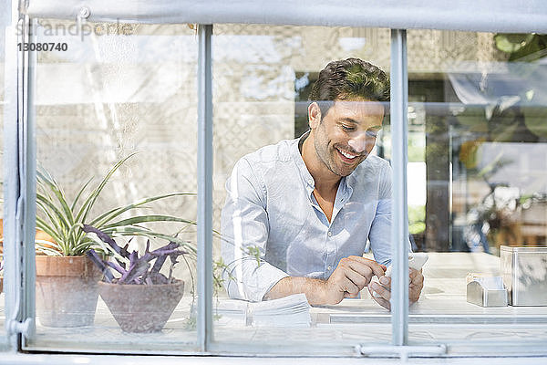 Glücklicher Mann mit Smartphone im Straßencafé durch Glasfenster gesehen