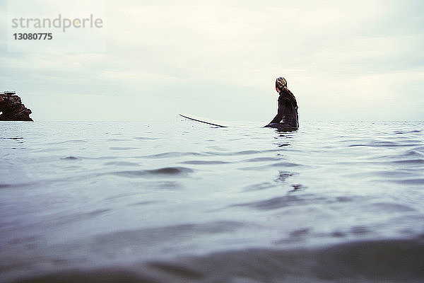 Seitenansicht einer Frau  die auf einem Surfbrett im Meer vor bewölktem Himmel sitzt