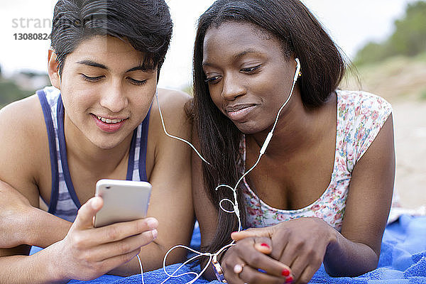 Junges Paar hört Musik über Smartphone  während es am Strand auf einer Decke liegt