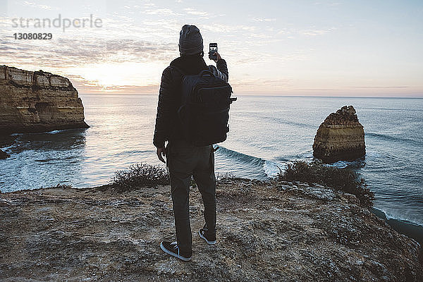 Rückansicht eines Mannes  der sich mit seinem Mobiltelefon selbstständig macht  während er bei Sonnenuntergang am Strand gegen den Himmel steht