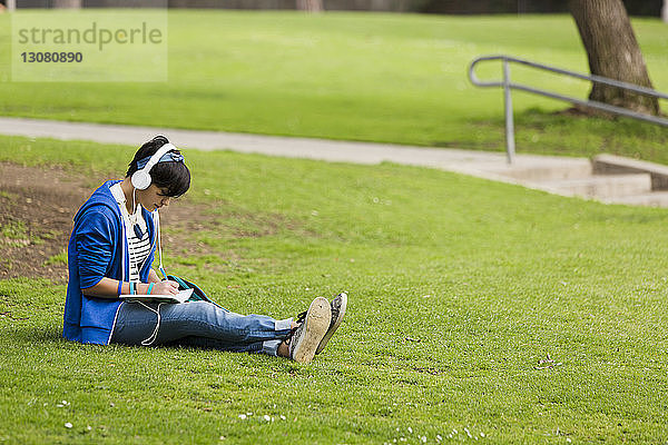Teenager-Mädchen in voller Länge hört Musik  während sie im Park an einem Tagebuch schreibt