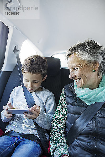 Glückliche ältere Frau sieht ihren Enkel an  der ein Smartphone im Auto benutzt