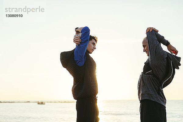 Seitenansicht von Vater und Sohn  die die Arme ausstrecken  während sie bei Sonnenuntergang am Strand gegen den klaren Himmel stehen