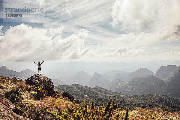 Fernansicht einer Wanderin  die mit ausgestreckten Armen auf einem Berg vor bewölktem Himmel steht