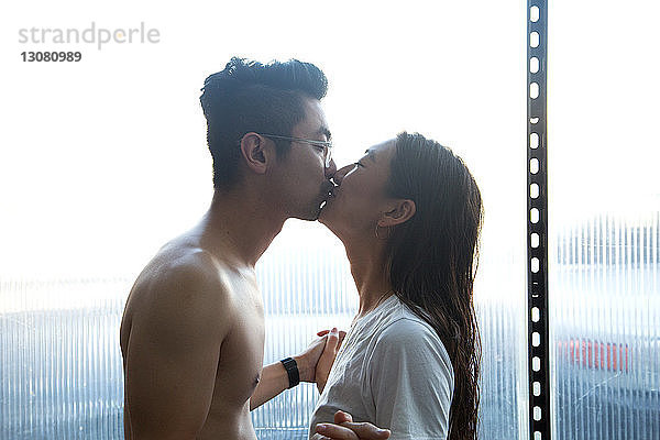 Seitenansicht eines romantischen Paares  das sich küsst  während es zu Hause am Fenster steht