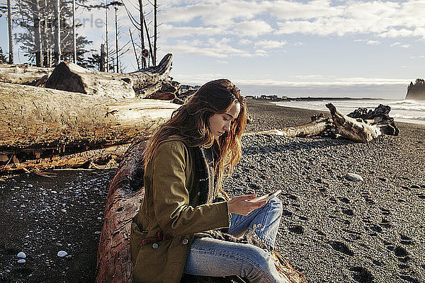 Seitenansicht einer Frau  die ein Smartphone benutzt  während sie am Strand sitzt