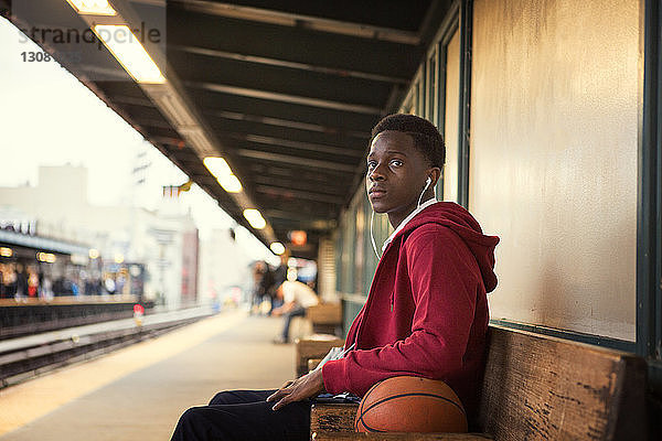 Seitenansicht eines Schülers  der Musik hört  während er am Bahnhof beim Basketball sitzt