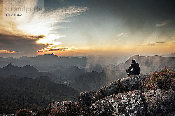 Rückansicht eines Mannes  der bei Sonnenuntergang auf einem Berg sitzt