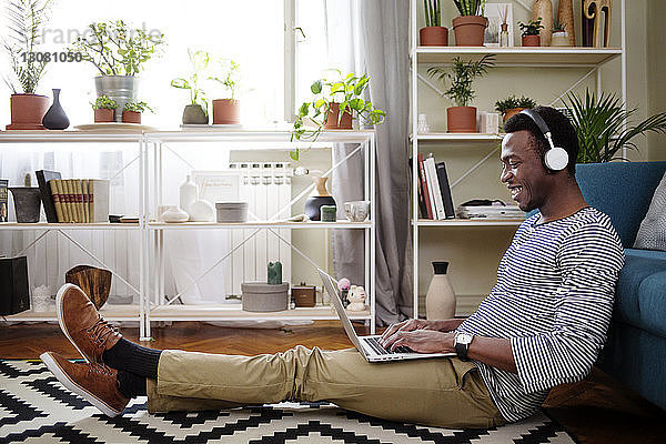 Seitenansicht eines glücklichen Mannes  der einen Laptop benutzt  während er im Wohnzimmer auf dem Boden sitzt