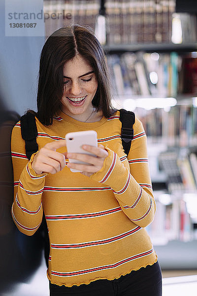 Lächelnde Frau benutzt Mobiltelefon  während sie in der Bibliothek steht