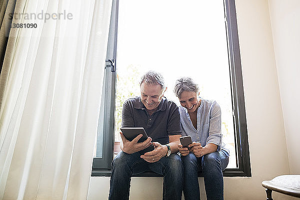Älteres Ehepaar nutzt Smartphone und Tablet-Computer  während es zu Hause auf dem Fensterbrett sitzt