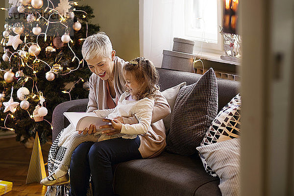 Großmutter hilft Mädchen beim Lesen von Büchern  während sie zu Hause auf dem Sofa sitzt