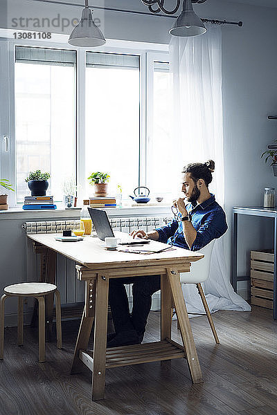Mann benutzt Laptop-Computer  während er zu Hause am Fenster sitzt