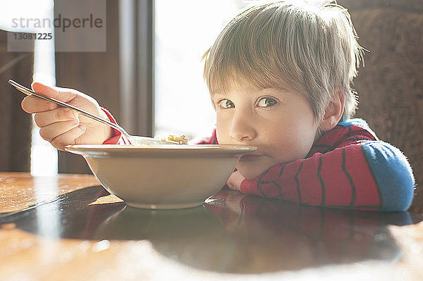 Porträt eines Jungen  der zu Hause Essen isst  während er sich an den Tisch lehnt