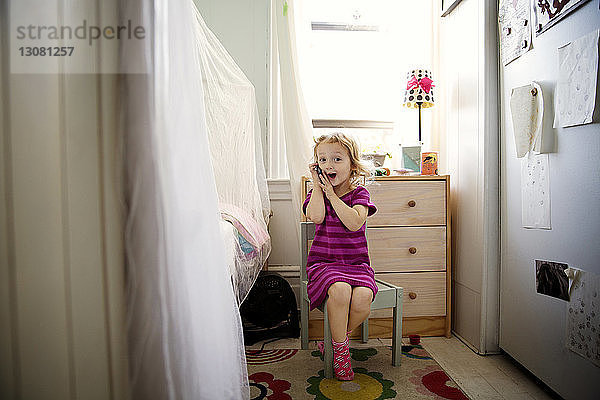 Süßes Mädchen spielt zu Hause auf einem Stuhl sitzend mit einem Mobiltelefon