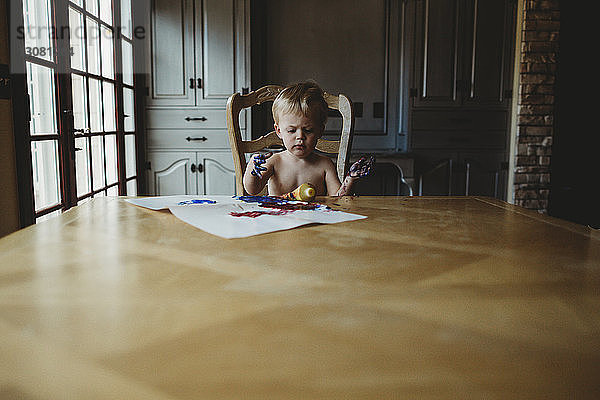 Kleiner Junge ohne Hemd mit schmutzigen Händen  der zu Hause am Holztisch sitzt