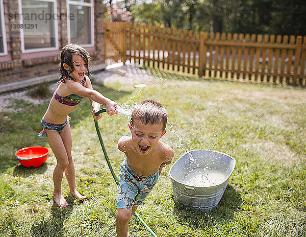 Hochwinkelansicht der verspielten Schwester  die im Hof durch einen Schlauch Wasser auf den hemdlosen Bruder spritzt