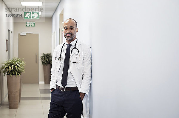 Porträt eines lächelnden Arztes mit an die Wand gelehnten Händen in Taschen im Krankenhaus