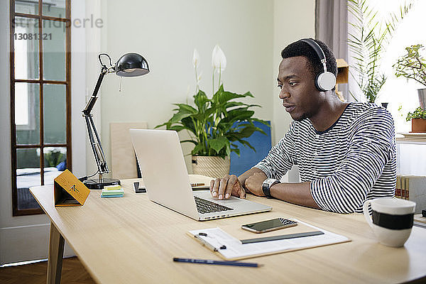 Junger Mann hört Musik  während er den Laptop im Heimbüro benutzt