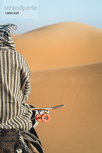 Rückansicht eines Mannes mit Skateboard  der bei Sonnenschein auf einem Kamel sitzt
