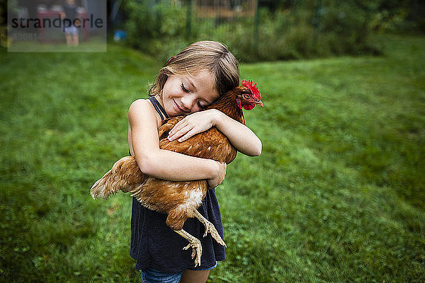 Lächelndes Mädchen mit geschlossenen Augen hält Henne  während sie im Hof steht