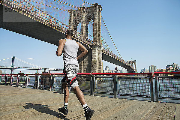 Rückansicht eines Mannes  der auf der Strandpromenade gegen die Brooklyn Bridge rennt