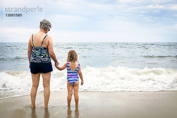 Rückansicht von Enkelin und Großmutter  die am Strand am Ufer stehen