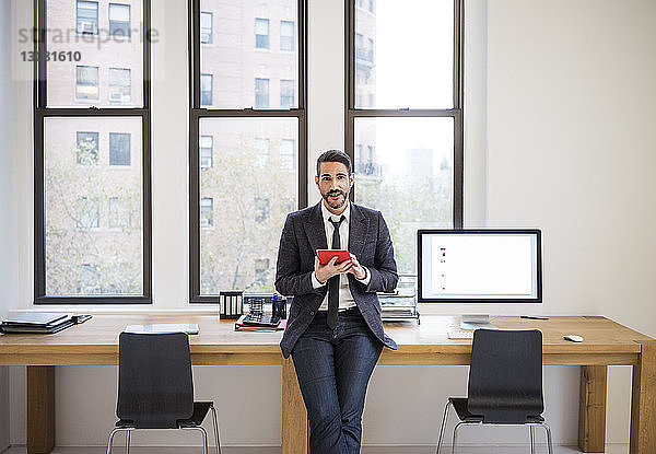 Porträt eines Geschäftsmannes  der einen Tablet-Computer benutzt  während er sich im Büro auf den Schreibtisch lehnt