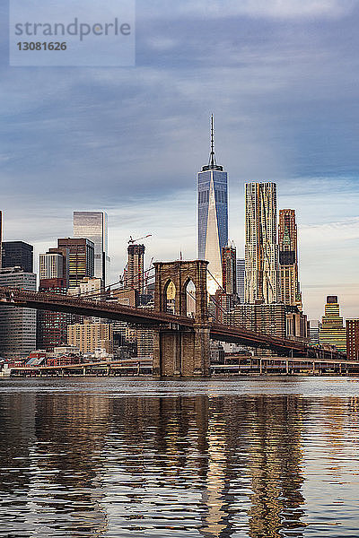 Brooklyn Bridge über den East River mit Stadtlandschaft im Hintergrund vor bewölktem Himmel