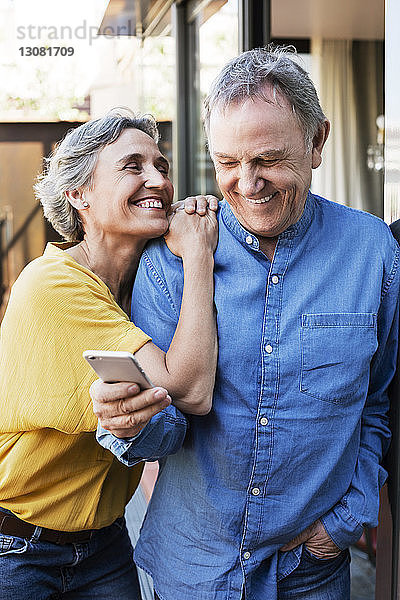 Fröhliche Frau sieht älteren Mann an  der ein Smartphone auf der Veranda benutzt