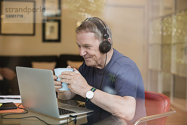 Glücklicher älterer Mann trinkt Kaffee  während er zu Hause über seinen Laptop Musik hört