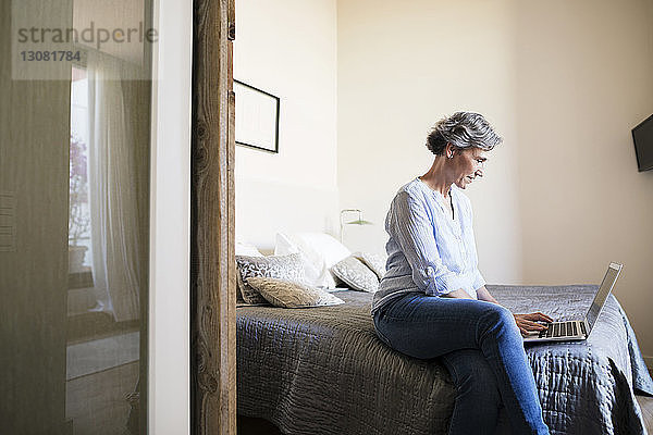 Seitenansicht einer reifen Frau  die einen Laptop benutzt  während sie zu Hause auf dem Bett sitzt