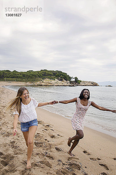 Fröhliche Freundinnen halten sich an den Händen  während sie am Strand gegen den Himmel rennen