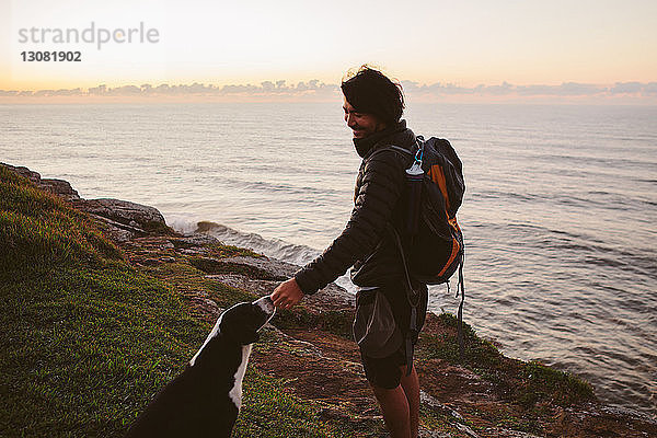 Seitenansicht eines männlichen Wanderers  der einen Hund berührt  während er auf einem Hügel am Meer steht