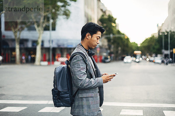 Seitenansicht eines Geschäftsmannes mit Rucksack  der ein Smartphone benutzt  während er auf der Straße steht