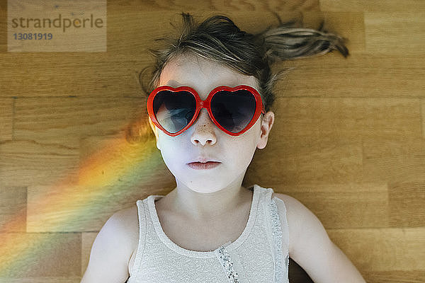 Draufsicht eines Mädchens  das eine herzförmige Sonnenbrille trägt und zu Hause auf einem Hartholzboden liegt