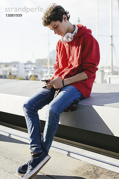 Teenager benutzt Smartphone  während er an einem sonnigen Tag auf einer Stützmauer sitzt