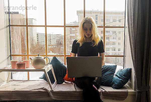 Frau benutzt Laptop-Computer auf einem Hocker am Fensterplatz in einer Nische zu Hause