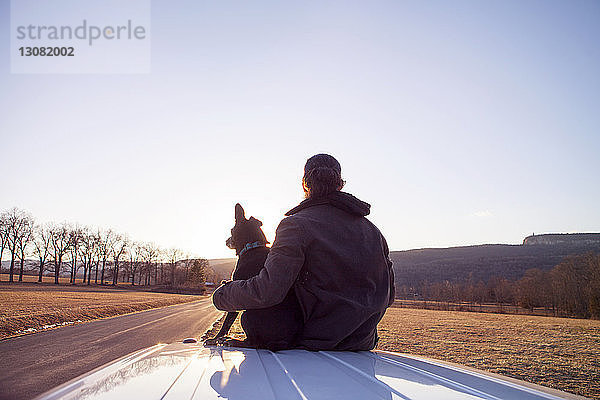 Rückansicht eines Mannes mit Hund auf dem Autodach bei klarem Himmel